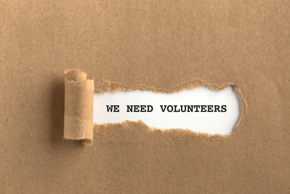 WE NEED VOLUNTEERS type-written behind craft paper section torn back over text