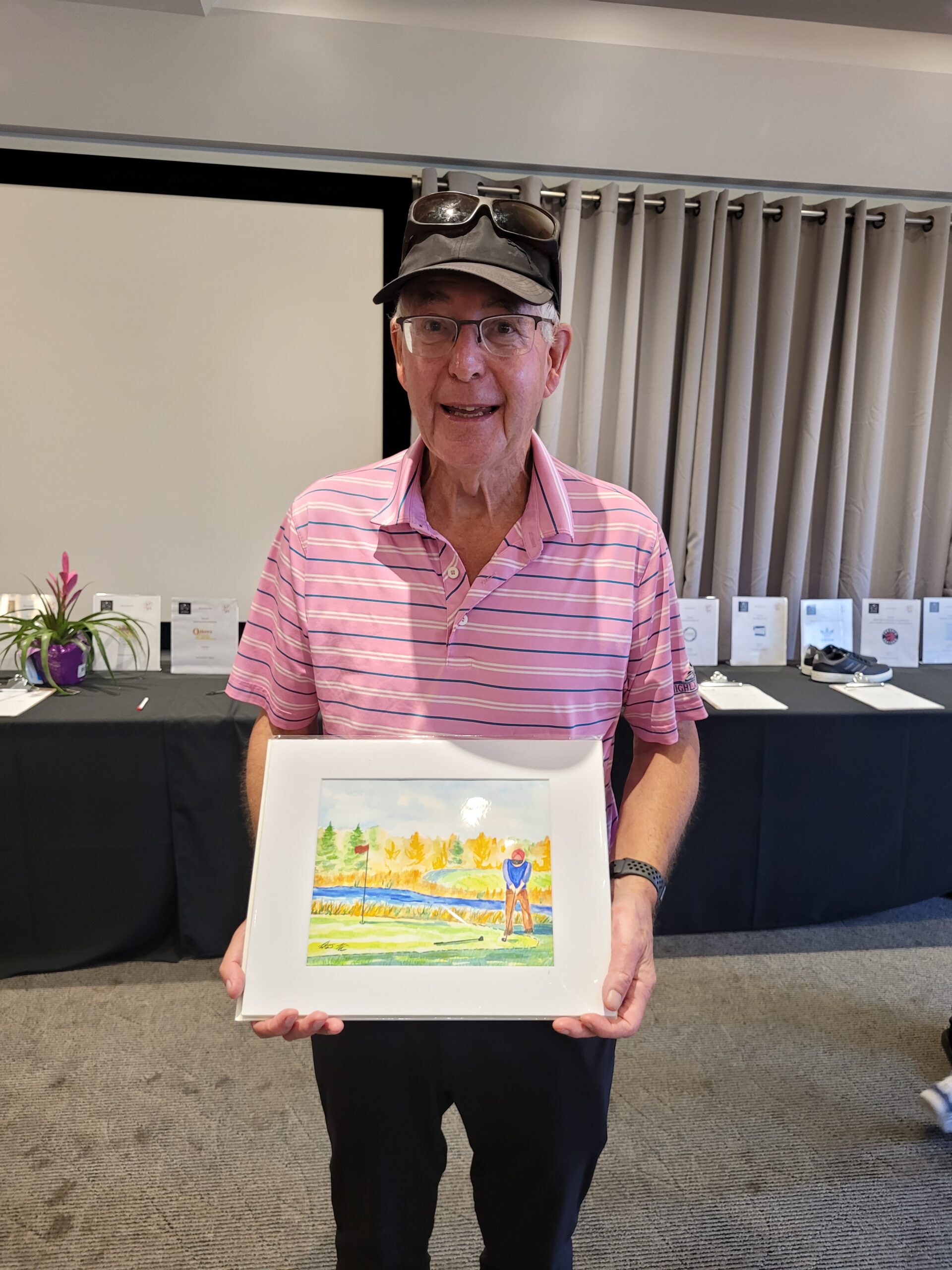 A smiling older man wearing a pink striped polo shirt and a black cap holds up a framed painting. Behind him, there's a black table covered with various items and a plant in a purple pot. Neutral-colored curtains are visible in the background.